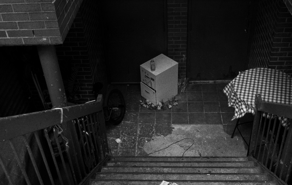 Black and white view from stairs into a tiled courtyard: a filing cabinet, bicycle, and checkered table among brick walls and deep shadow.
